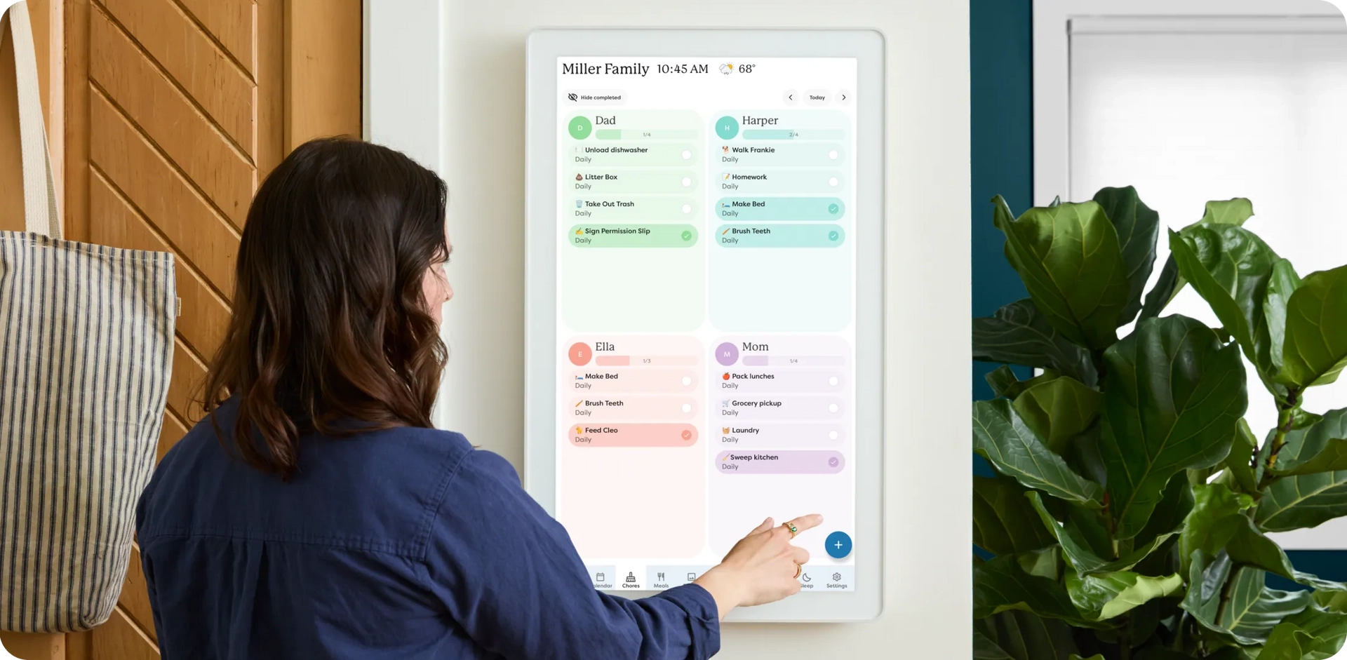 Women interacting with a Skylight Calendar Max wall-mounted digital display in portrait mode, showing a family task organizer with customizable sections for each member