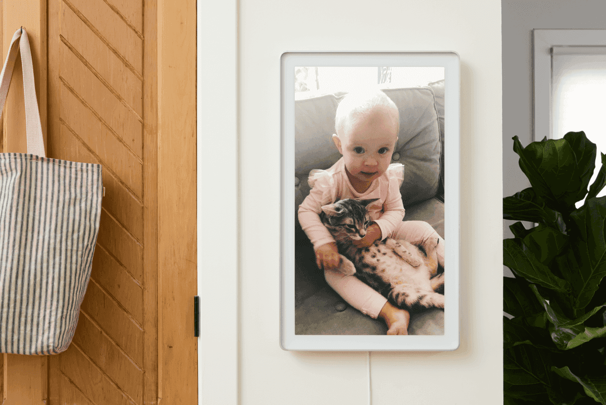 A photo frame mounted on a wall displays an image of a smiling baby sitting on a couch, holding a relaxed tabby kitten